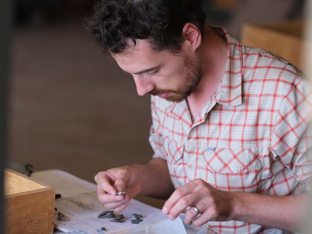 A man wearing a short sleeve, plaid shirt bends over a table and works on small, rock-life artifacts.