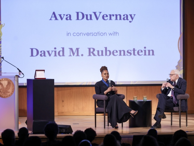 A woman and a man sit in two chairs on a stage speaking into mics. 