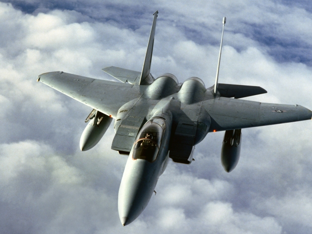 A fighter plane with narrow nose and wide wings in a light gray color flies above the clouds.