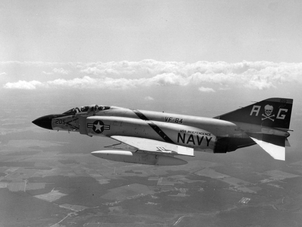 Black and white image of a small plane with military painting flies over clouds and open fields. 