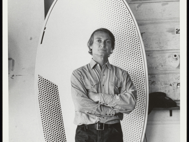 Black and white image of a man standing with his arms crossed in front of a painted surfboard