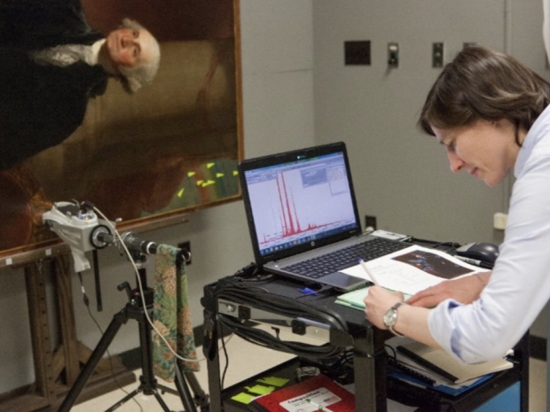 A person leans over a table writing in a notebook with a computer and camera set up to analyze a piece of art featuring George Washington.