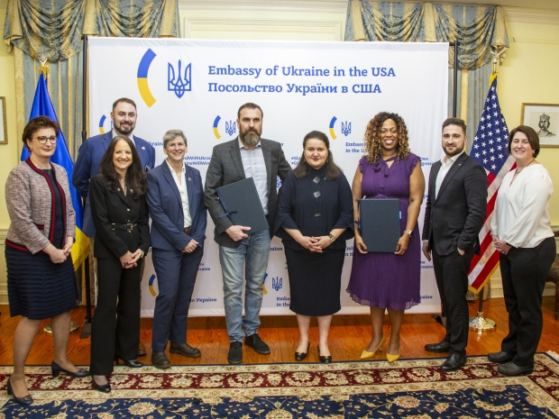 A group of people, smiling toward camera, stand against a white backdrop in large room.