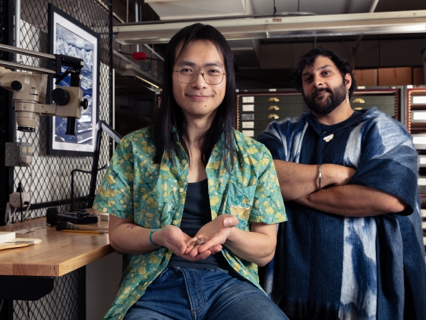 A man holds a small fossil in his hands as another stands behind him with his arms crossed.