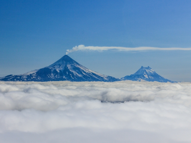 Two volcanoes, one with a stream of smoke coming out of the top, seen above a plume of clouds