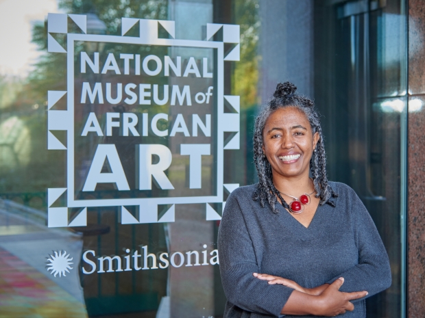 Woman stands with arms gently crossed, smiling, wearing a grey top and red necklace.
