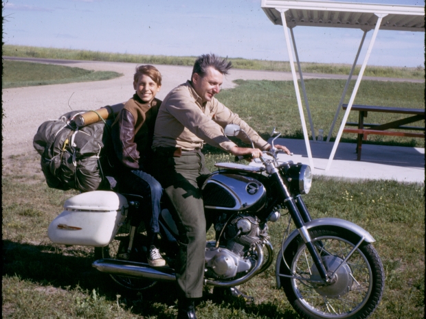 Vintage picture of a man siting on a black motorcycle and a small boy sitting on seat behind him.