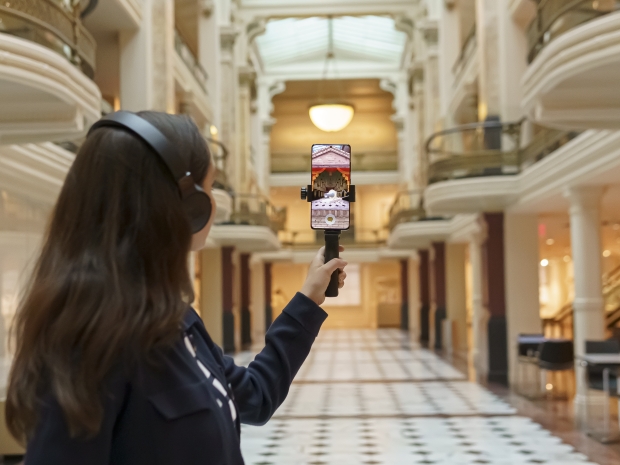 Girl with brown hair and black headphones holds a phone up in a large space, projecting an augmented reality graphic.
