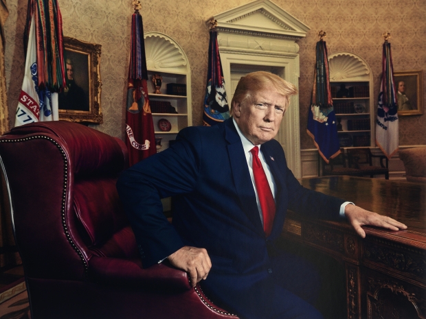 Man in black suit and red tie sits at a dark wooden desk in the Oval Office.