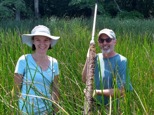 A man and a woman wearing short sleeve shirts and outdoors attire stand in field of tall green gas holding large stick with soil 