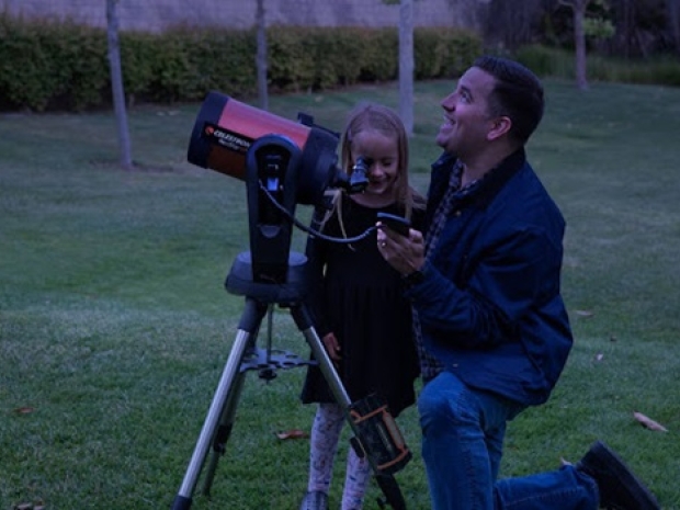 An older man kneels next to a telescope and young girl looking at the sun. 