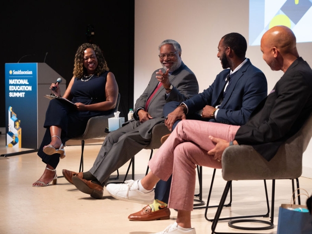 A panel of four adults are smiling in conversation seated on a stage