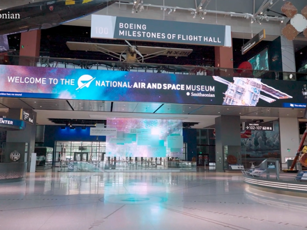 Interior of museum lobby with large white floor, a screen at ceiling height, and large aircraft on display.