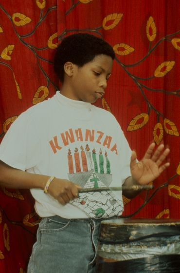 Young boy wearing a white shirt with Kwanzaa written on it drums with a stick and his hand.