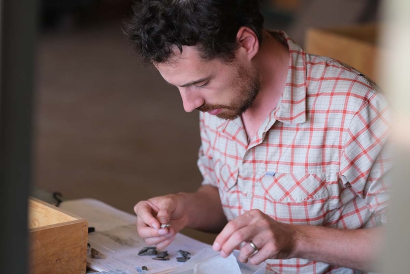 A man wearing a short sleeve, plaid shirt bends over a table and works on small, rock-life artifacts.