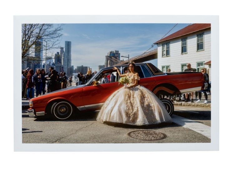 Color photograph of a girl in a large white ballgown standing in front of a red car propped up on hydraulic back tires.