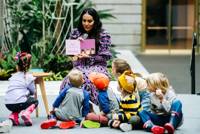 A woman in a long dress sits on a stool reading a book to a group of children.