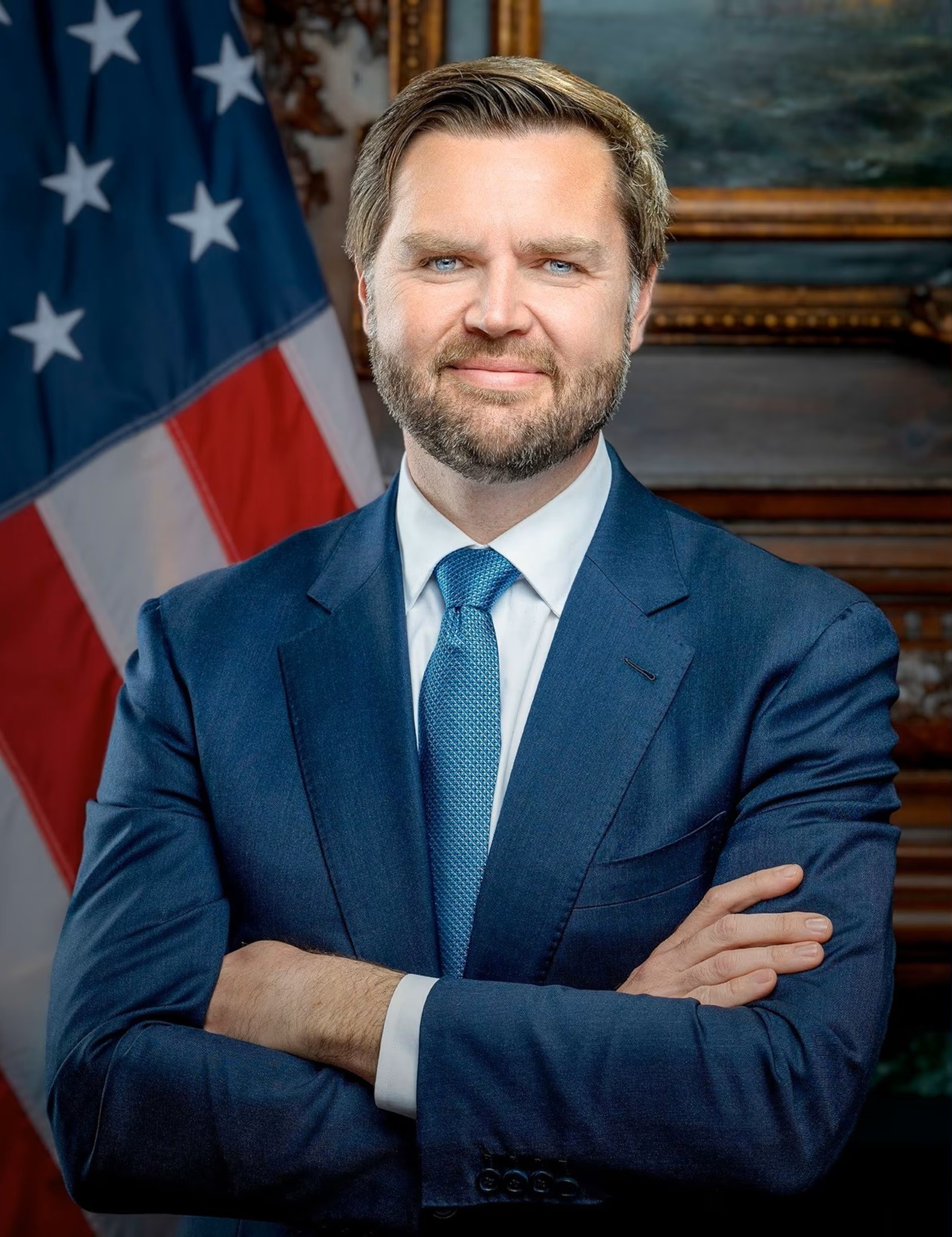 Photograph of Vice President JD Vance, man wearing a suit and standing with his arms folded. The American flag is in the background.